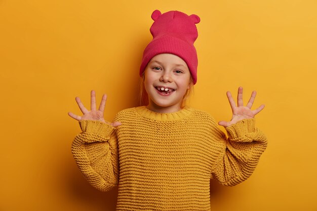 Be positive and keep smiling. Glad adorable European child raises hands and shows palms, expresses good emotions, plays with someone, dressed in knitted sweater, isolated on yellow wall