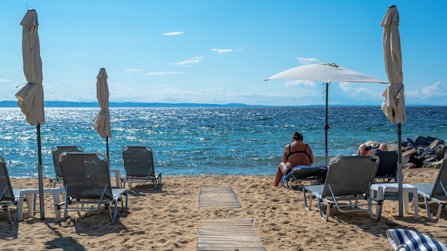 Beach on Aegean sea coast with umbrellas and sunbeds, resting couple, rocks near the water in Nikiti, Greece