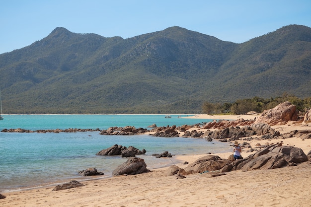 Beach covered in rocks surrounded by the sea and hills covered in forests under sunlight