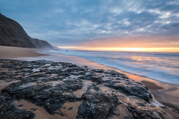 Beach surrounded by rocks and the sea under a cloudy sky during a beautiful sunset