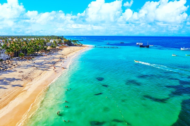해변 휴가 및 여행 배경입니다. 짚 우산, 야자수, 보트가 있는 아름다운 대서양 열대 해변의 공중 무인 항공기 보기. 바바로 해변, 푼타 카나, 도미니카 공화국.