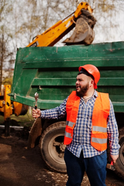 Beard worker man suit construction worker in safety orange helmet against dump truck with hammer and adjustable wrench at hand