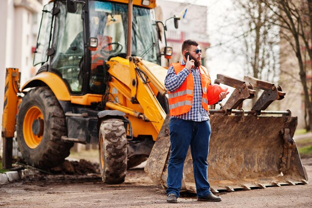 Beard worker man suit construction worker in safety orange helmet sunglasses against tractor with mobile phone at hand