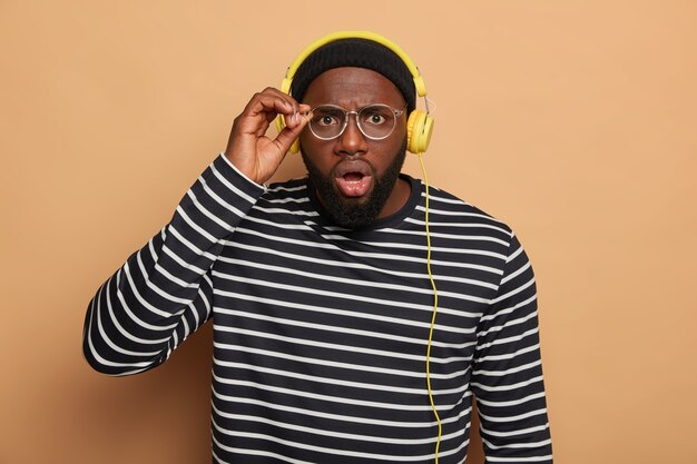 Bearded adult man looks scrupulously through round glasses, has shocked reaction