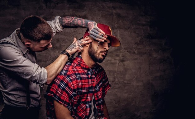 Foto gratuita barbuto afroamericano hipster con una camicia di pile e un berretto che si fa tagliare i capelli da un parrucchiere professionista vecchio stile fa il taglio di capelli.