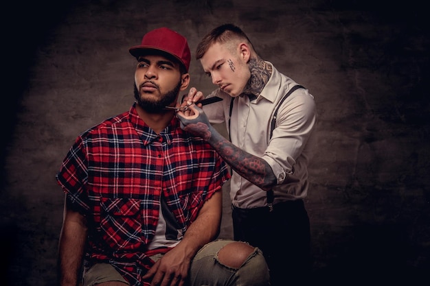 Bearded African American hipster guy in a fleece shirt and cap getting a haircut by an old-fashioned professional hairdresser does haircut.
