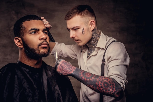 Barbuto afroamericano hipster che si fa tagliare i capelli da un parrucchiere professionista tatuato vecchio stile fa il taglio di capelli.