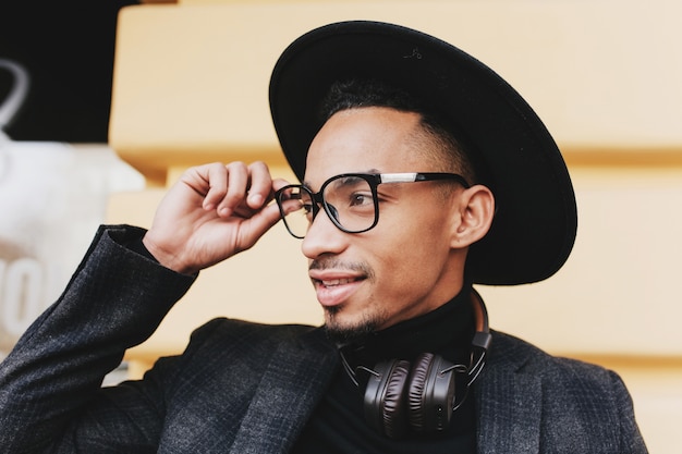 Bearded african man with short haircut touching his glasses. Close-up outdoor photo of black boy in hat and music headphones.