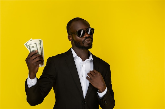 Bearded afroamerican guy is holding dollars in one hand, wearing sunglasses and black suit 