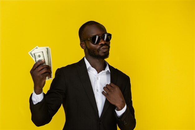 Bearded afroamerican guy is holding dollars in one hand, wearing sunglasses and black suit 