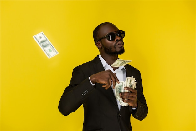 Bearded afroamerican guy is throwing out dollars from one hand, wearing sunglasses and black suit 