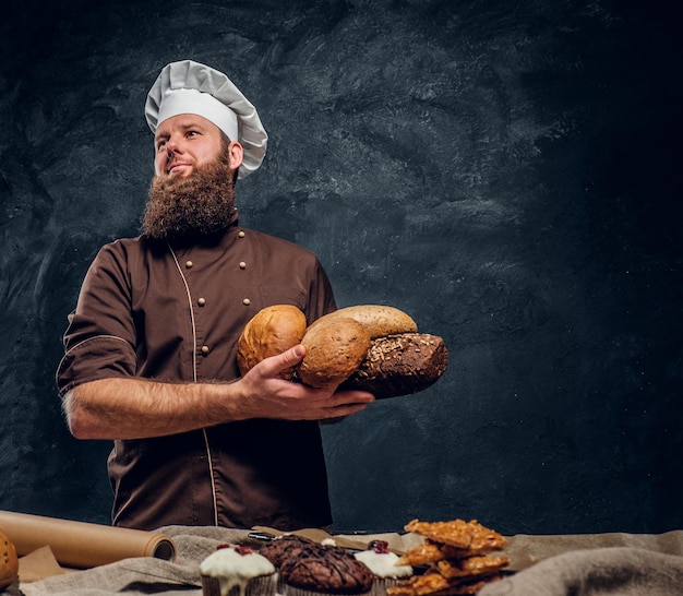 A bearded baker wearing a uniform holding fresh bread, standing next to a table, decorated with delicious bread loaves, baguettes and muffins in a dark studio