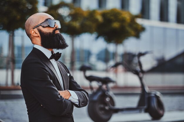 Бородатый лысый бизнесмен стоит, скрестив руки, возле своего электроскутера на улице.