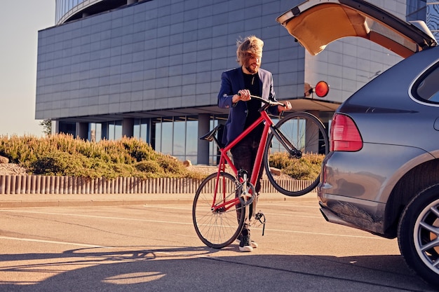 Bearded blond male with long hair holds fixed bicycle near the car with open trunk.