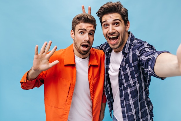 L'uomo barbuto brunet in camicia a scacchi prende selfie e mette le orecchie da coniglio al suo amico. il tipo con la giacca arancione non vuole fare foto.