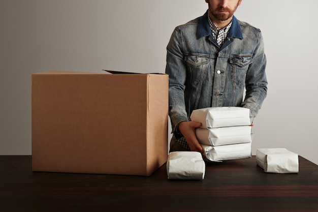 Uomo barbuto brutale in giacca da lavoro jeans mette pacchetti ermetici sigillati vuoti all'interno di una grande scatola di carta cartone sulla tavola di legno. consegna speciale