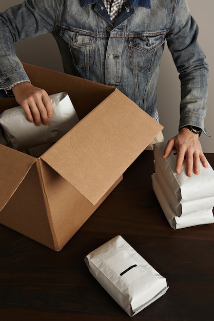 Uomo barbuto brutale in giacca da lavoro jeans mette pacchetti ermetici sigillati vuoti all'interno di una grande scatola di carta cartone sulla tavola di legno. consegna speciale
