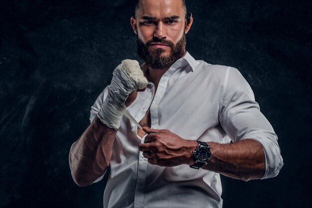 Bearded brutal man in white shirt is wearing protection on his fist before fight at dark studio.