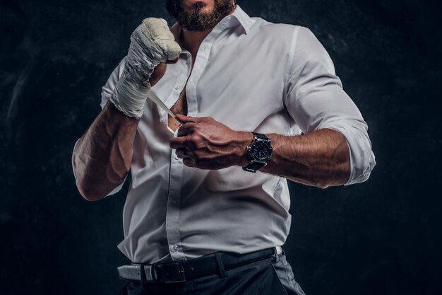 白いシャツを着たひげを生やした残忍な男は、暗いスタジオで戦う前に彼の拳に保護を身に着けています。