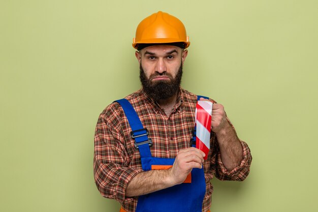 Bearded builder man in construction uniform and safety helmet holding adhesive tape looking at camera confused standing over green background