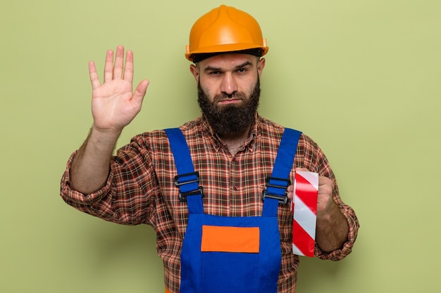 Bearded builder man in construction uniform and safety helmet holding adhesive tape looking with serious face making stop gesture with hand