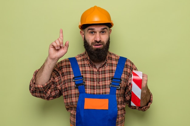 Bearded builder man in construction uniform and safety helmet holding adhesive tape looking with smile on face showing index finger having new idea