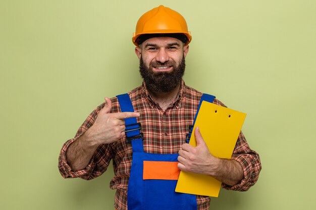 Bearded builder man in construction uniform and safety helmet holding clipboard pointing with index finger at it happy and confident smiling