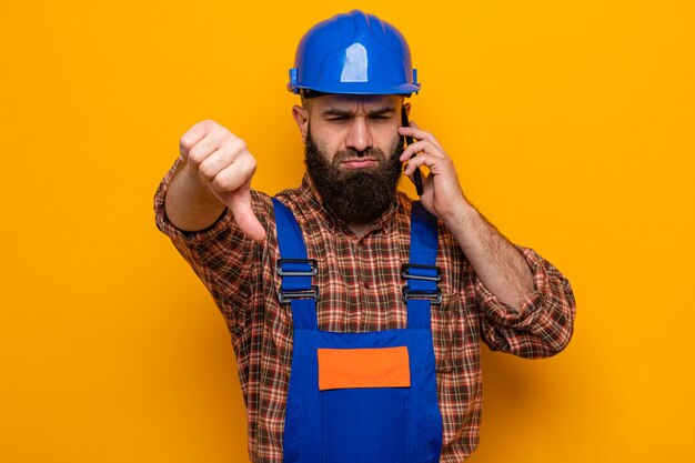 Bearded builder man in construction uniform and safety helmet looking at camera displeased showing thumbs down talking on mobile phone standing over orange background