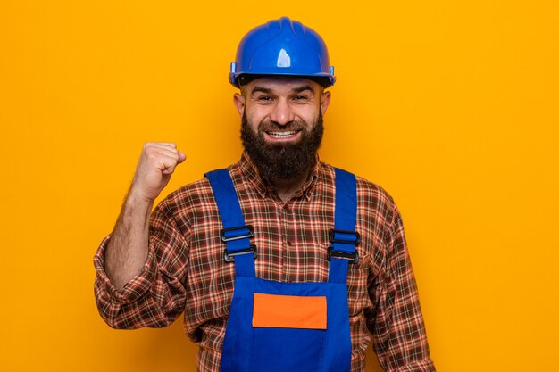 Бородатый строитель в строительной форме и защитном шлеме смотрит в камеру счастливым и возбужденным, сжимая кулак, весело улыбаясь, стоя на оранжевом фоне