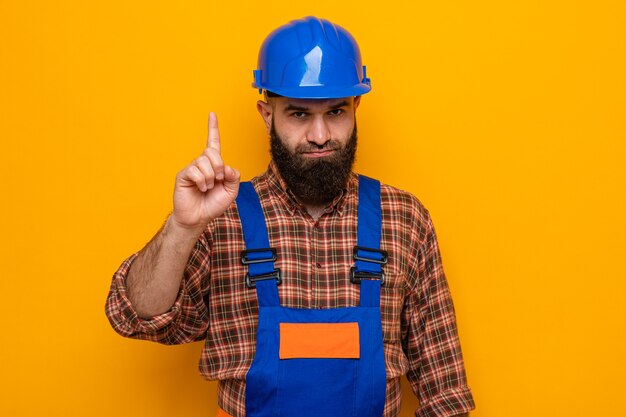 Bearded builder man in construction uniform and safety helmet looking at camera with serious face showing index finger warning gesture standing over orange background