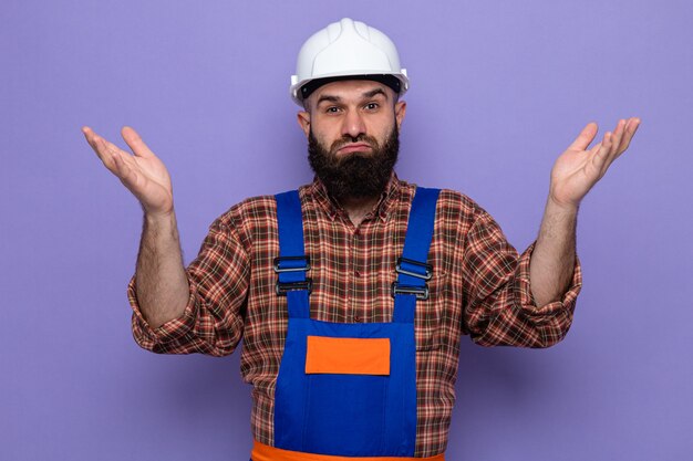 Bearded builder man in construction uniform and safety helmet looking at canera confused raising arms having no answer