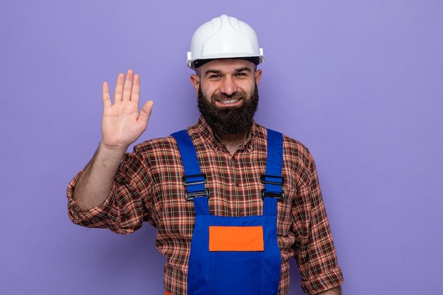 Bearded builder man in construction uniform and safety helmet looking smiling cheerfully showing fifth with palm