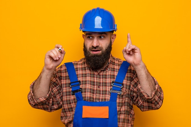 Free photo bearded builder man in construction uniform and safety helmet looking surprised and happy writing with pen in air