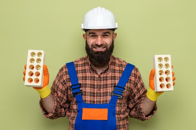 Bearded builder man in construction uniform and safety helmet wearing rubber gloves holding bricks looking at camera smiling cheerfully happy and positive standing over green background