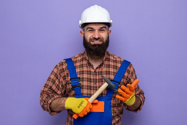 Bearded builder man in construction uniform and safety helmet wearing rubber gloves holding a hammer looking at camera smiling cheerfully happy and positive standing over purple background