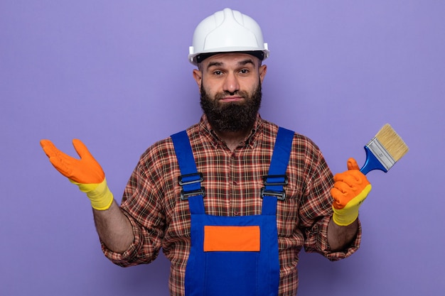 Бородатый мужчина-строитель в строительной форме и защитном шлеме в резиновых перчатках держит кисть с краской и смотрит в камеру в замешательстве, разводя руки в стороны, стоя на фиолетовом фоне
