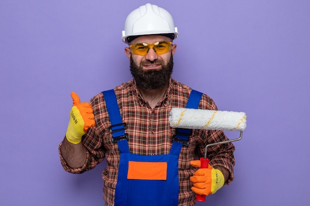 Bearded builder man in construction uniform and safety helmet wearing rubber gloves holding paint roller looking at camera smiling showing thumbs up standing over purple background