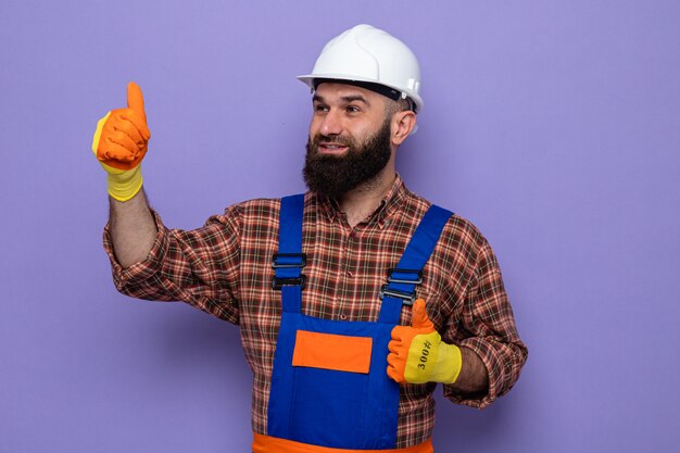 건설 유니폼을 입은 수염 난 건축업자 남자와 고무 장갑을 끼고 안전모를 쓰고 행복하고 쾌활한 엄지손가락을 옆으로 바라보고 있습니다.