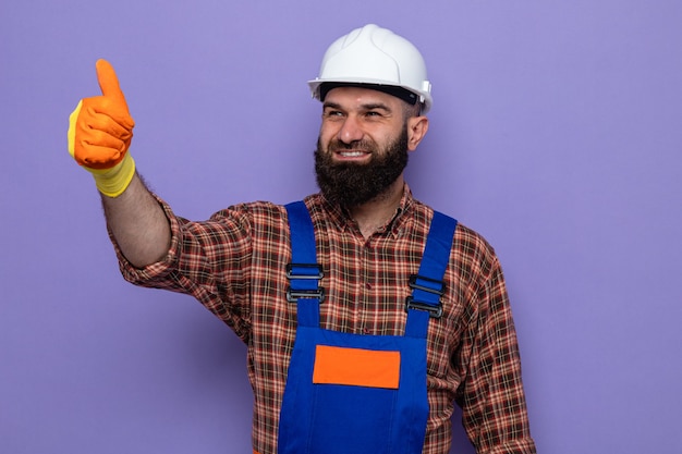 建設制服と安全ヘルメットのひげを生やしたビルダーの男は、紫色の背景の上に立って親指を元気に笑顔で脇を見てゴム手袋を着用