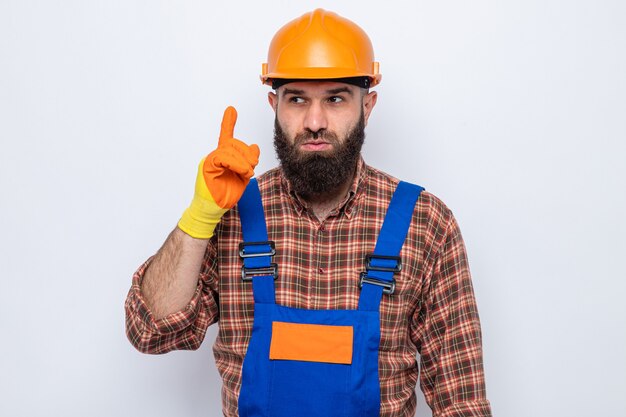 Bearded builder man in construction uniform and safety helmet wearing rubber gloves looking aside surprised showing index finger having new idea