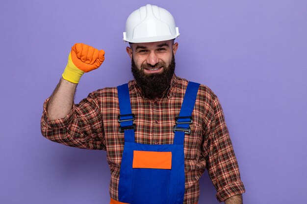 Bearded builder man in construction uniform and safety helmet wearing rubber gloves looking at camera happy and confident smiling raising fist like a winner standing over purple background