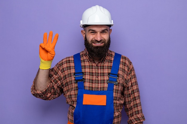 建設制服と安全ヘルメットのひげを生やしたビルダーの男は、指で3番目を元気に笑顔で見ているゴム手袋を着用しています