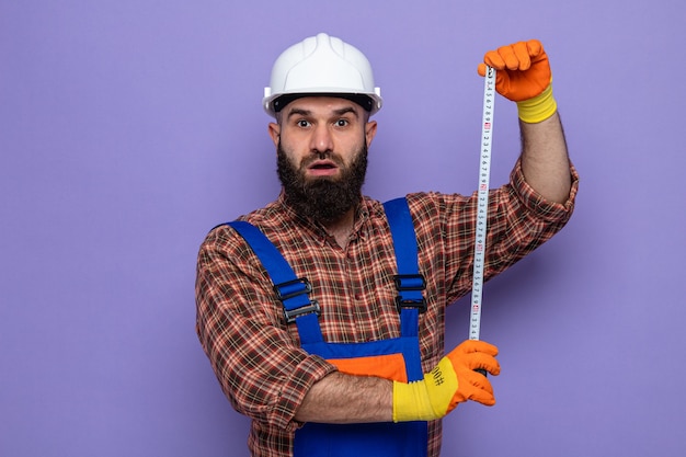 Bearded builder man in construction uniform and safety helmet wearing rubber gloves looking surprised working using measure tape