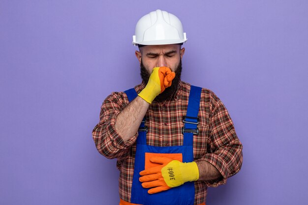 紫色の背景の上に立っている拳で体調不良の咳をしているゴム手袋を身に着けている建設制服と安全ヘルメットのひげを生やしたビルダーの男