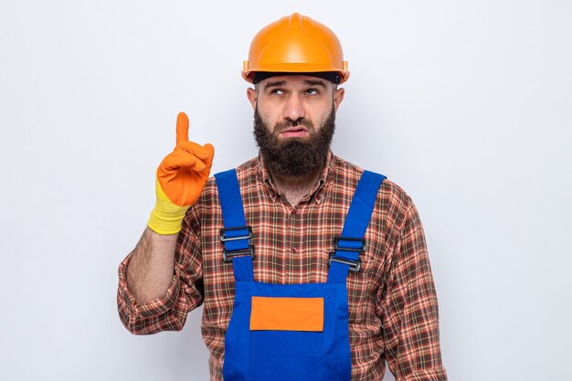 Бородатый строитель в строительной форме и защитном шлеме в резиновых перчатках озадаченно смотрит вверх, показывая указательный палец, стоящий на белом фоне