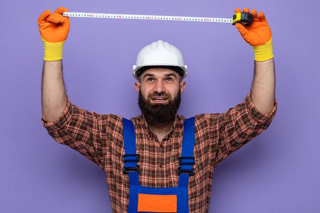 Bearded builder man in construction uniform and safety helmet wearing rubber gloves looking up with confident expression working using measure tape standing over purple background