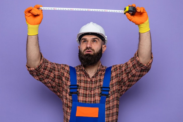 Bearded builder man in construction uniform and safety helmet wearing rubber gloves looking up with confident expression working using measure tape