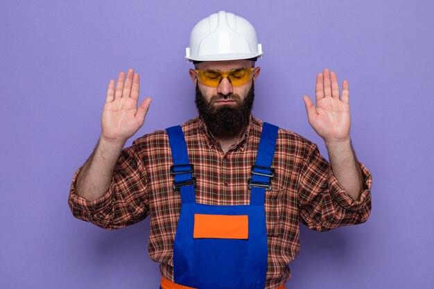 보라색 배경 위에 자신감 있게 팔을 들고 있는 노란색 안전 안경을 쓴 건설 유니폼과 안전 헬멧을 쓴 수염 난 건축업자