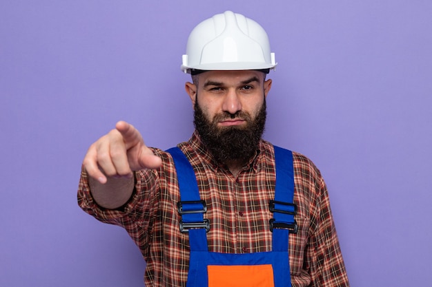 Бесплатное фото Бородатый мужчина-строитель в строительной форме и защитном шлеме, указывая указательным пальцем на камеру, глядя с серьезным лицом, стоящим на фиолетовом фоне
