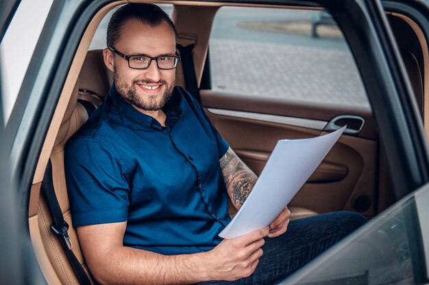 Bearded business male in eyeglasses with tattoo on his arm sits on a back seat of a car.